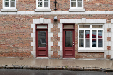 Two vintage half glass, half-wooden red colored antique doors. The facade of the brown and red brick building has beige block trim. There are three windows on the second floor and one on the main.