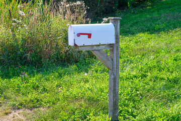 A white aluminum metal retro-style postal box on a wooden post. There's a small red mail flag on the side of the rectangle shaped container. The rural letterbox is surrounded by green grass and trees.