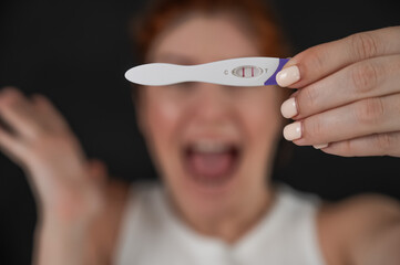 Blurred portrait of upset caucasian woman holding positive rapid pregnancy test on black background. 