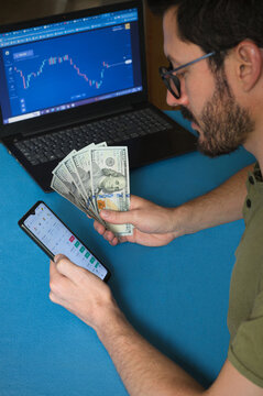 Operador, Trader de Forex. Hombre joven haciendo Trading con laptop, tel&eacute;fono m&oacute;vil y d&oacute;lares sobre superficie azul. Mercado de divisas. Binance, Cripto, Bitcoin. 