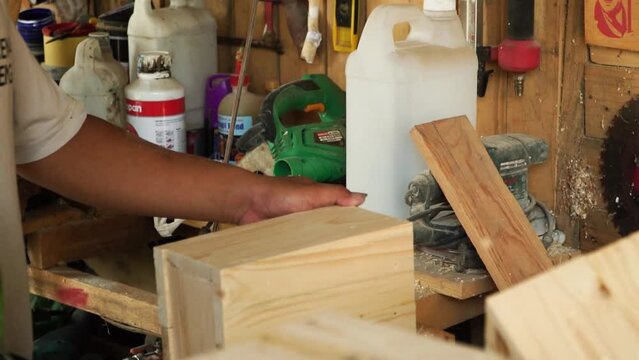 Close up of a carpenter working in a simple wood workshop. The workshop is full of piles of wood, circular saws, nailer guns, planing machines, drilling machines, wood glue and even wood paint