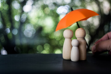 Family of wooden dolls are hiding under a orange umbrella, protecting wooden peg dolls with copy...