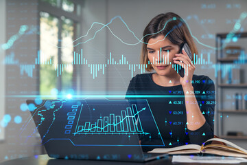 Pensive attractive beautiful businesswoman in working on laptop, talking phone, office workplace in background. Forex graph and charts hologram icons. Concept of market trading and research.