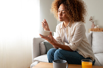 Young relaxed pensive African American woman eating healthy breakfast sitting on the couch. Copy space. - obrazy, fototapety, plakaty