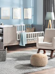 Contemporary baby's room with pastel blue walls, showcasing a gallery mockup of three blank frames, a sleek wooden crib featuring a grey cushion, an adjacent beige armchair, and a plush grey area rug.