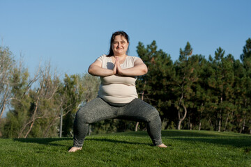 Overweight middle-aged woman doing weight loss exercises in park. Happy fat woman doing plie squats standing barefoot on grass. Healthy lifestyle, body positivity, Pilates outdoors in summer