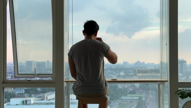 While drinking coffee, Asian man sits in solitude on a balcony, his demeanor exuding a profound sense of sadness. The atmosphere is heavy with melancholy.