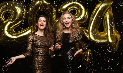 Two young ladies drinking champagne. Image of girls with balloons isolated on black background, having fun at New Year's 2021 Eve Party.