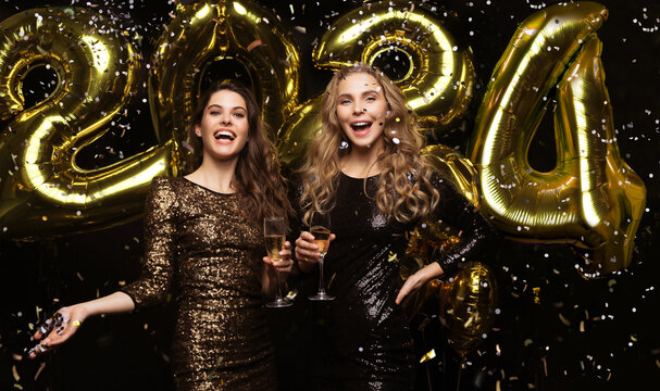 Two young ladies drinking champagne. Image of girls with balloons isolated on black background, having fun at New Year's 2021 Eve Party.
