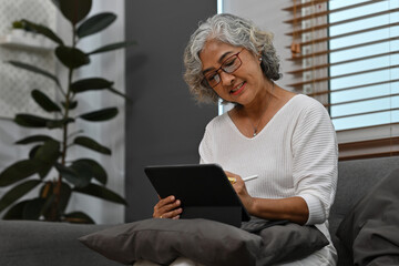 A smiling Senior Asian woman sits and relax on the couch in the living room while browsing wireless internet on a tablet, elderly with a technology concept