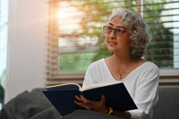 A smiling Senior Asian woman relaxes on the couch in the living room while reading a book, or literature, enjoying free time on the weekend. Elderly people's lifestyles and leisure activity concepts