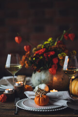 Elegant seasonal fall table setting with floral composition in a pumpkin vase as a centrepiece. White wine, burning candles, romantic cozy atmosphere of family dinner, private autumn wedding
