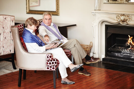 Senior couple, relax and reading by fire place on holiday, retirement or vacation together at hotel or resort. Man and woman sitting with magazine or paper for news, deals or activities at house