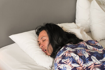Long haired asian man is sleeping with prone position and his mouth open on a bed with a white pillow. Unusual sleep posture at the hotel during holiday vacation. Rest after a long journey
