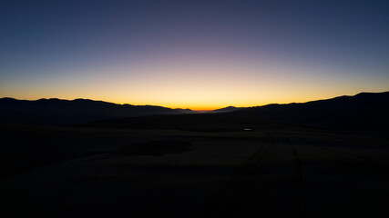 Dawn in the mountains. The violet-orange color of the sky. The red rays of the sun come out from behind the peaks. A light haze in the gorge. Traces of the road and puddles are visible. Drone view