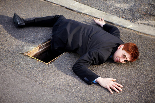 Portrait, accident and an unlucky business man on a street in the city after falling into a hole. Mistake, stuck or clumsy and a ginger corporate professional on the road in a suit with a problem