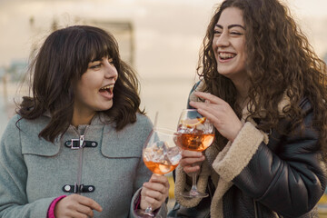 Joyful moment between two friends with drinks - Two young women share a joyful moment, laughing and toasting with spritz cocktails, radiating happiness and friendship.
