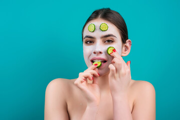 Naked young woman with clay mask and cucumbers on face, isolated on blue. Beautiful model applying cosmetic cream. Facial treatment. Topless woman with bare shoulders applying face cream.