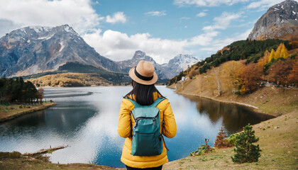 Rear view of a stylish girl, with a backpack, a hat and a yellow jacket, looking at the view of the mountains and the lake while relaxing in the autumn nature. Travel concept.