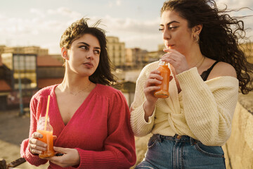 Two young women relish the warm glow of the setting sun on a city rooftop, each with a bottle of...