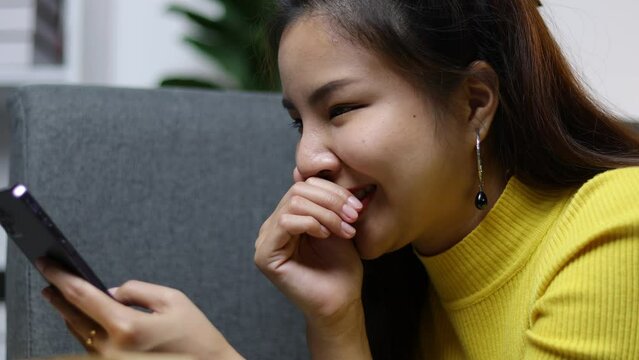 Young Asian woman happily playing with mobile phone and laughing in living room at home.