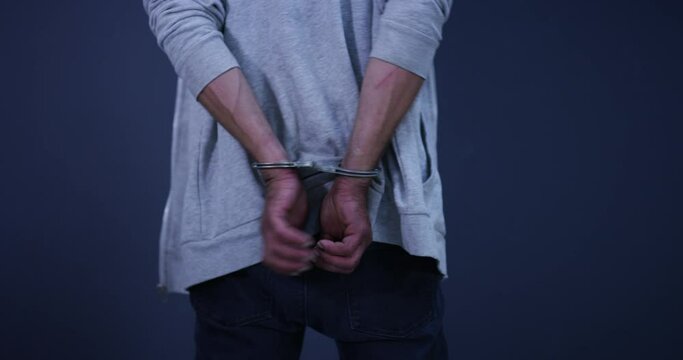 Hands, cuffs and closeup of man prisoner in studio for illegal crime, danger or corruption. Arrest, surrender and zoom of male criminal or suspect in jail for theft, scam or fraud by blue background.