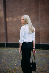 A beautiful smiling slender woman with long blond hair, in a black long dress and a white shirt, stands on a city street, against the backdrop of a granite building wall.