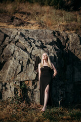 A slender, positive, sexy aged woman with long blond hair in a black long stylish dress stands near the picturesque rocks on a bright sunny summer day.