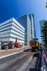 BASEL, SWITZERLAND - July 11 2023: Roche Towers - skyscrapers in the Swiss city of Basel. Road service workers shoveling new asphalt during construction and patching of a road. - obrazy, fototapety, plakaty