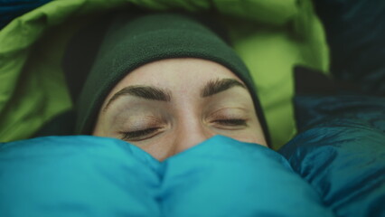 Woman wake up in camp tent sleeping bag. Close up portrait of tourist girl opening eyes in cold morning. Female traveler wilderness vacation, tourism and camping in outdoor trip.