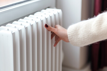 Woman in sweater warming her hands at heating radiator close up - obrazy, fototapety, plakaty