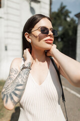 Urban fashion summer beautiful woman model with sunglasses with tattoo on arm in stylish beige dress walks in sunny summer day. Pretty girl
