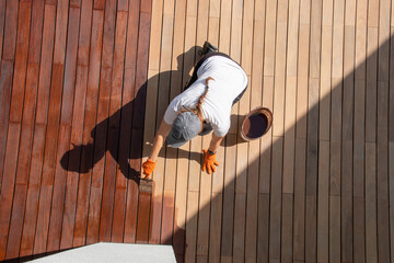 Woman applying brown wood protection oil on decking boards with paint brush, terrace renovation top view - obrazy, fototapety, plakaty