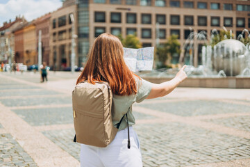 Attractive young female tourist is exploring new city. Redhead woman pointing finger and holding a paper map in a warm sunny day. Urban lifestyle concept. Traveler