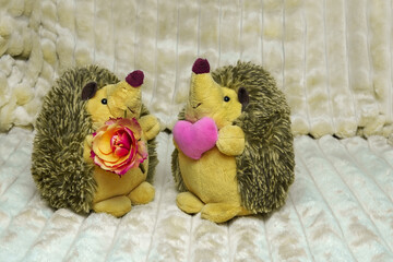 Two toy hedgehogs with yellow bellies and gray needles, one has a rose flower, the other has a large toy heart in his hands. They confess their love
