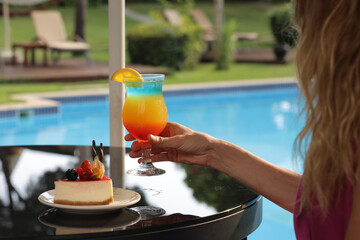 A woman with juice in her hand sitting at the bar by the pool, cocktail and cake on the table at...