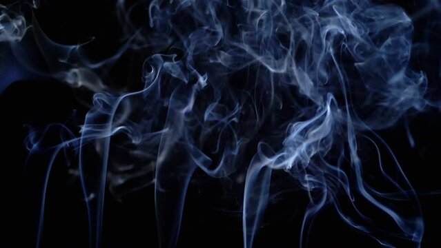 Thin Curls of Blue Smoke Rise Up, Fill Empty Space on a Black Background. Incense sticks. Abstract shapes. Texture. Beautiful clouds of smoke swirl. Floating fog, flowing smoke. Blurred motion. Light.