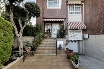Frontal image of the façade of a single-family home with stairs leading to the porch, a ramp down to the garage, various pots and a garden with ornamental trees and hedges