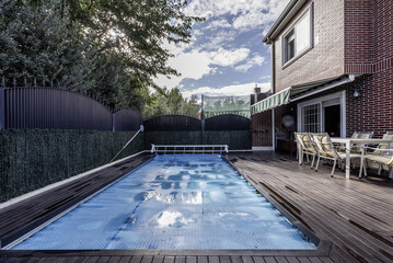 Backyard of a single-family home with a swimming pool covered by retractable protective canvas on a floor covered with acacia wood slats