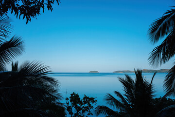 Silhouetted palms frame a calm, azure expanse, where distant islands nestle under a deep blue sky.