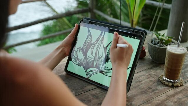 Female illustrator hands draw on tablet at cafe with ocean view, palm trees. Freelancer working outdoor on the terrace at the seaside. Pretty woman drink cappuccino. Close-up, back view.