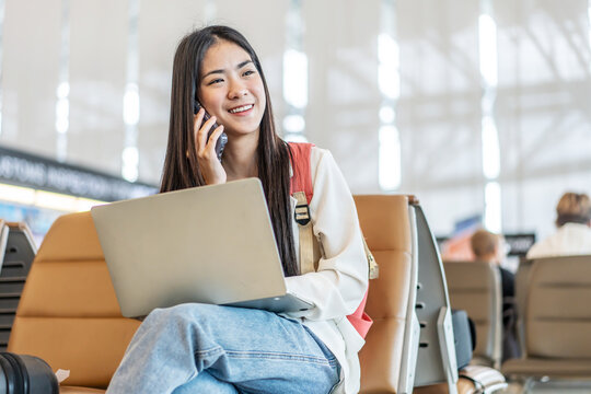Smiling young asian woman sitting in waiting room and talking over smartphone with friend or family and using laptop. Beautiful female traveller talking on cell phone at airport lounge.