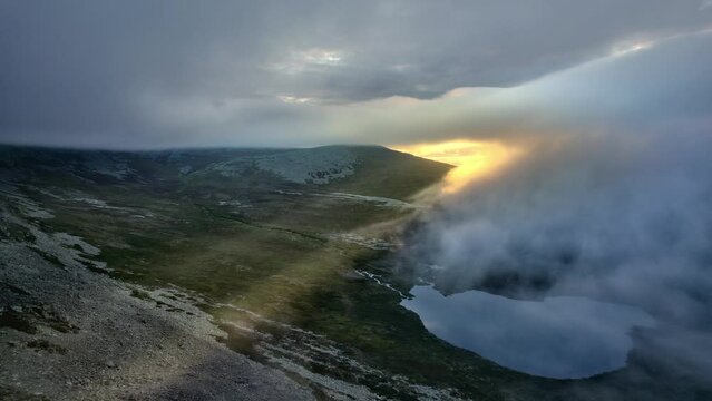 Aerial drone shot of sunrise in the mountains, fiery yellow sun rays breaking through the fog and white clouds. Flying over tundra landscape with mountain river and lake, Swedish wilderness