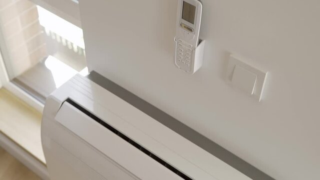 The woman presses the power button on the remote control of the air conditioner mounted on the wall. The home air conditioner turns on, opens the air outlet and starts pumping cold air into the room.
