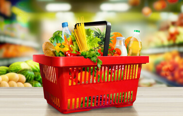 Groceries in red shopping basket on wooden table in suppermarket aisle in background