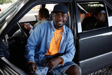 Young smiling male hip hop dancer in black cap, denim jacket and shorts sitting on front seat of car and looking at camera against his buddies - obrazy, fototapety, plakaty