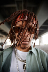 African American guy with dreadlocks looking at camera through sunglasses while standing in urban environment under bridge - obrazy, fototapety, plakaty