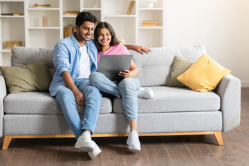 Happy Indian couple on sofa, using laptop computer together, free space