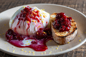 White mozzarella cheese served with raspberry jam and a slice of whole grain bread on a plate, closeup
