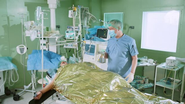 A professional mature surgeon in a robe and mask advises and soothes the patient under local anaesthesia before a serious operation in the surgeoning room. Doctor talking an African-American patient.